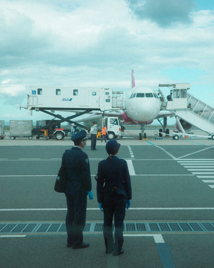 Two crew members approach a parked airplane at an airport, with a jet bridge connected.